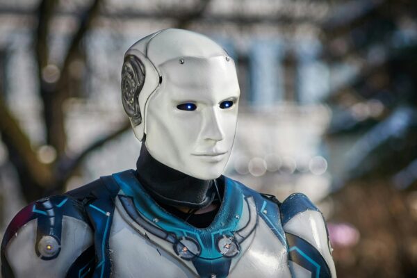 Close-up of a humanoid robot with glowing blue eyes, wearing futuristic white and blue armour, set against a blurred outdoor background.