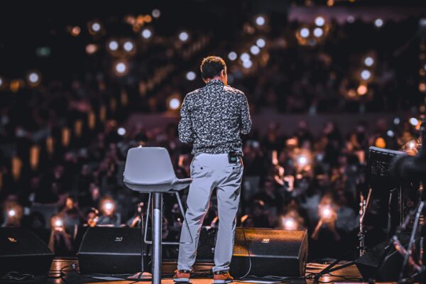 Male speaker on stage facing a large audience with lights glowing from phones, wearing a printed shirt and using a clip-on microphone, next to a modern grey stool.