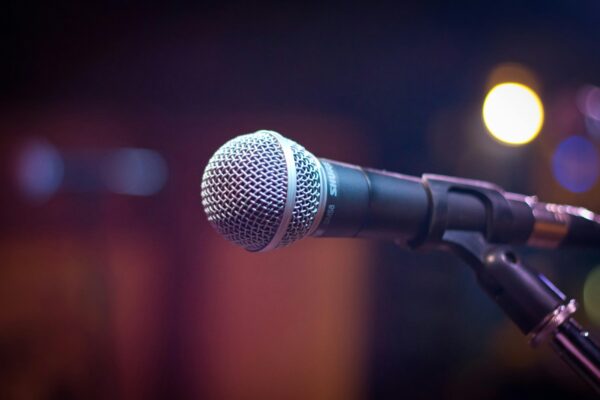Close-up of a handheld microphone on a stand, set against a blurred background with colourful stage lighting.